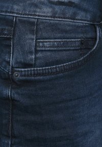 Los pantalones vaqueros de denim azul oscuro tienen un bolsillo trasero, costuras visibles y un botón negro en la cinturilla. Textura suave y mínimo desgaste.