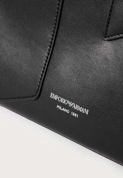 Κοντινή λήψη μαύρης δερμάτινης τσάντας Emporio Armani με ανάγλυφο λογότυπο και κείμενο "MILANO 1981" σε λευκό.