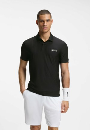 Fiatal férfi fekete BOSS pólóban, fehér BOSS rövidnadrágban és fehér csuklópántban, egyik kezét a zsebében tartva áll.