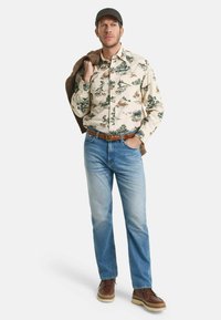 Blumengemusterte Button-Up-Bluse in Creme, blaue Jeans, braune Stiefel und eine Kappe; hält einen braunen Pullover über der Schulter.