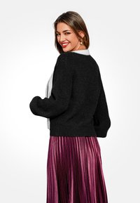 Cardigan nero lavorato a maglia con design cropped, caratterizzato da dettagli a coste e maniche ampie, abbinato a una gonna rosa lucida e plissettata.