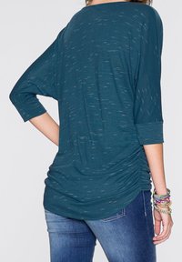 Haut ample couleur sarcelle avec manches trois-quarts, tissu texturé et côtés froncés. Associé à un jean en denim bleu et des bracelets colorés.