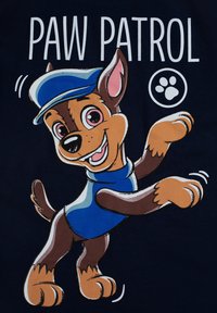 Personaggio di cane in stile cartone animato con pelo marrone, che indossa un'uniforme blu e un cappello, su uno sfondo blu navy scuro, con il testo "PAW PATROL" sopra.