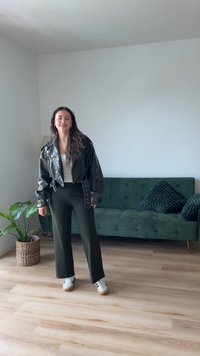 Veste de biker en cuir noir sur un haut blanc, associée à un pantalon large côtelé vert olive et des baskets blanches avec des semelles en caoutchouc. Canapé vert et plante en arrière-plan.