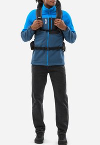 Blue fleece jacket with a zip, black adjustable straps, gray pants, and black hiking boots. The jacket features contrasting zippers and logos.