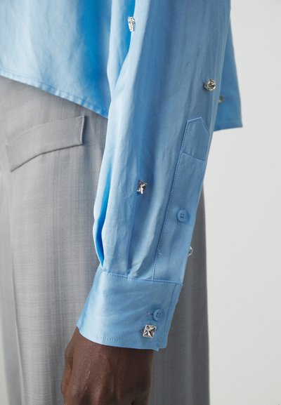 Cămașă bleu deschis cu guler ascuțit și butoni la manșete, având detalii din metal argintiu pe mânecă, asortată cu pantaloni gri texturați.