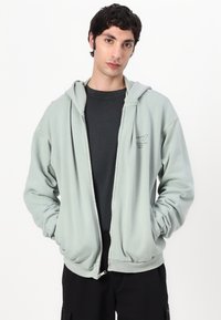 Felpa leggera verde chiaro con zip, dotata di cappuccio regolabile con cordino, realizzata in tessuto morbido, con tasche laterali e dettaglio del logo sul petto.