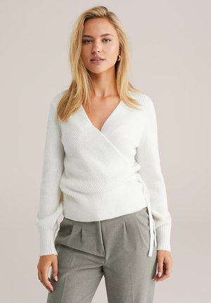Suéter blanco de punto acanalado con mangas largas y detalle atado en el costado, combinado con pantalones gris ajustados. Textura suave, diseño de escote en V profundo.