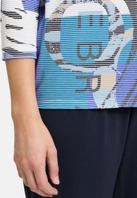 Mehrfarbiges Oberteil mit horizontalen blauen und weißen Streifen, das abstrakte Muster und ein rundes Grafikdesign in Blau und Beige zeigt. Schwarze Hose sichtbar.