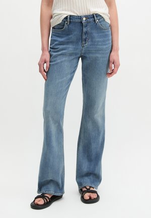 Frau trägt hellblaue High-Waist Schlagjeans, ein weißes geripptes Top und schwarze Riemchensandalen und steht vor einem weißen Hintergrund.