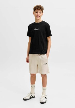 Nastolatek ubrany w czarny T-shirt z nadrukiem, beżowe szorty z kieszeniami, kremowe skarpetki z napisem i białe trampki, stojący na jednolitym tle.