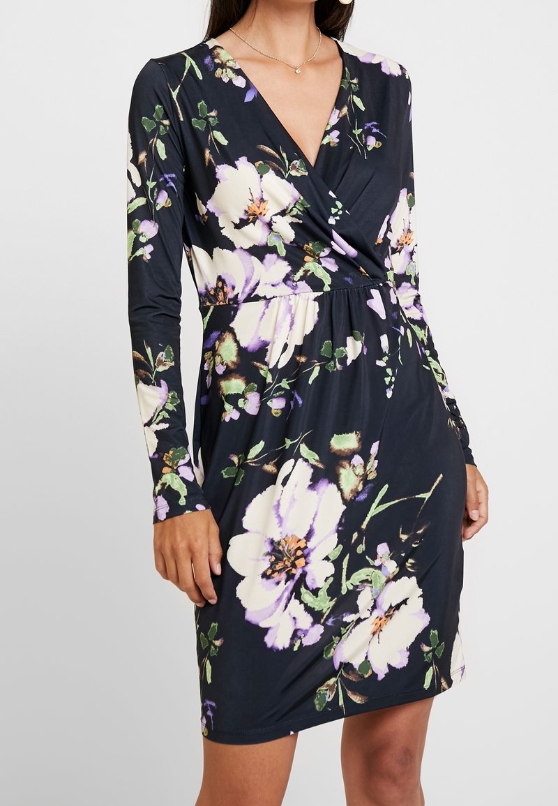 Femme portant une robe noire à manches longues avec un grand imprimé floral blanc et violet et un corsage croisé en V.