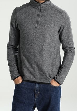 Mand iført en grå langærmet pullover med kvart lynlås, hænder i lommerne, kombineret med mørkeblå jeans mod en ensfarvet baggrund.