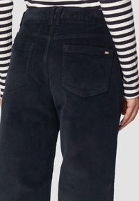 Donkere marineblauwe corduroy broek met een gestructureerd oppervlak, voorzien van twee achterzakken en een hoge taille. Gestreepte lange mouwen top zichtbaar.