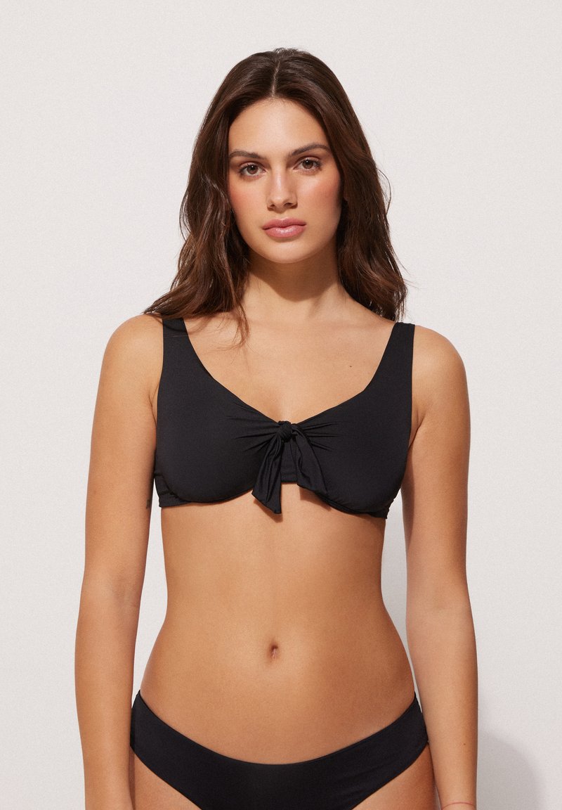 Schwarzer Bikini-Oberteil aus glattem Stoff, mit einem Knoten-Detail vorne und breiten Trägern, ergänzt durch ein einfaches Design mit klassischer Silhouette.