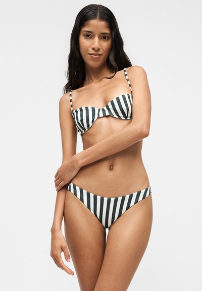 Femme aux longs cheveux foncés portant un bikini rayé vertical noir et blanc avec de fines bretelles sur un fond clair uni.