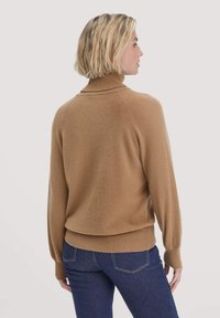 Strickpullover mit Rollkragen in Camel-Farbe, mit gerippten Bündchen und Saum. Das Material wirkt weich und hat eine lockere Passform. Kombiniert mit dunkelblauen Jeans.