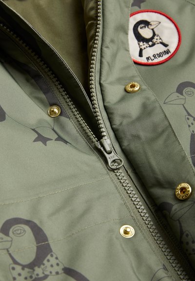 Žalia striukė su užtrauktuku priekyje, aukso spalvos detalėmis ir siuvinėtu pingvino lipduku; audinys yra minkštos tekstūros su subtiliu raštu.