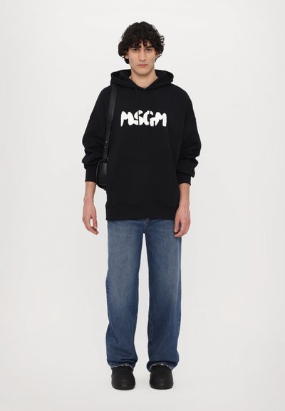 Felpa nera con cappuccio e logo grafico bianco sulla parte anteriore, abbinata a jeans larghi blu chiaro. Il modello porta anche una piccola borsa nera.