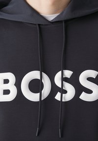Gros plan d'un sweat à capuche noir avec capuche grise et cordons noirs, présentant de grandes lettres blanches texturées formant le mot BOSS sur la poitrine.