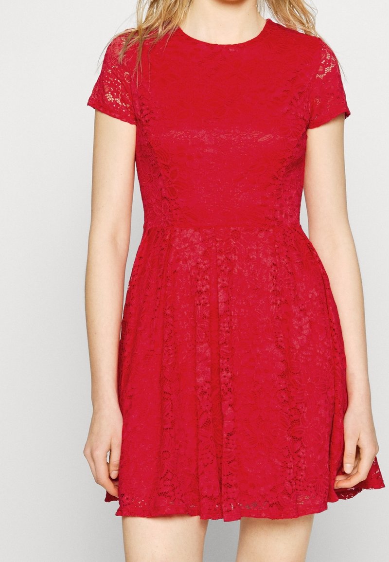 Femme portant une robe en dentelle rouge à manches courtes, taille cintrée et jupe évasée, debout devant un fond uni clair.