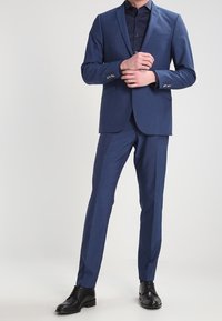 Marineblauer zweiteiliger Anzug aus strukturiertem Stoff, bestehend aus einem Blazer mit Reverskragen und schmal geschnittenen Hosen, kombiniert mit schwarzen eleganten Schuhen.