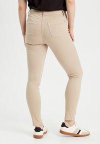 Jean skinny beige avec une texture lisse, cinq poches et une taille mi-haute, associés à des baskets blanches avec des accents noirs.