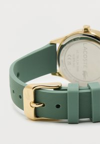 Zelený silikonový pásek na hodinky s jemným vzorem, zlatá kovová spona a zlatý pouzdro hodinek. Na pásku je viditelné logo Lacoste.