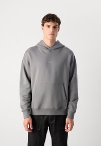 Grå hettegenser med lommer foran, myk tekstur, ribbede mansjetter og en liten logo på brystet. Brukt med mørke jeans.