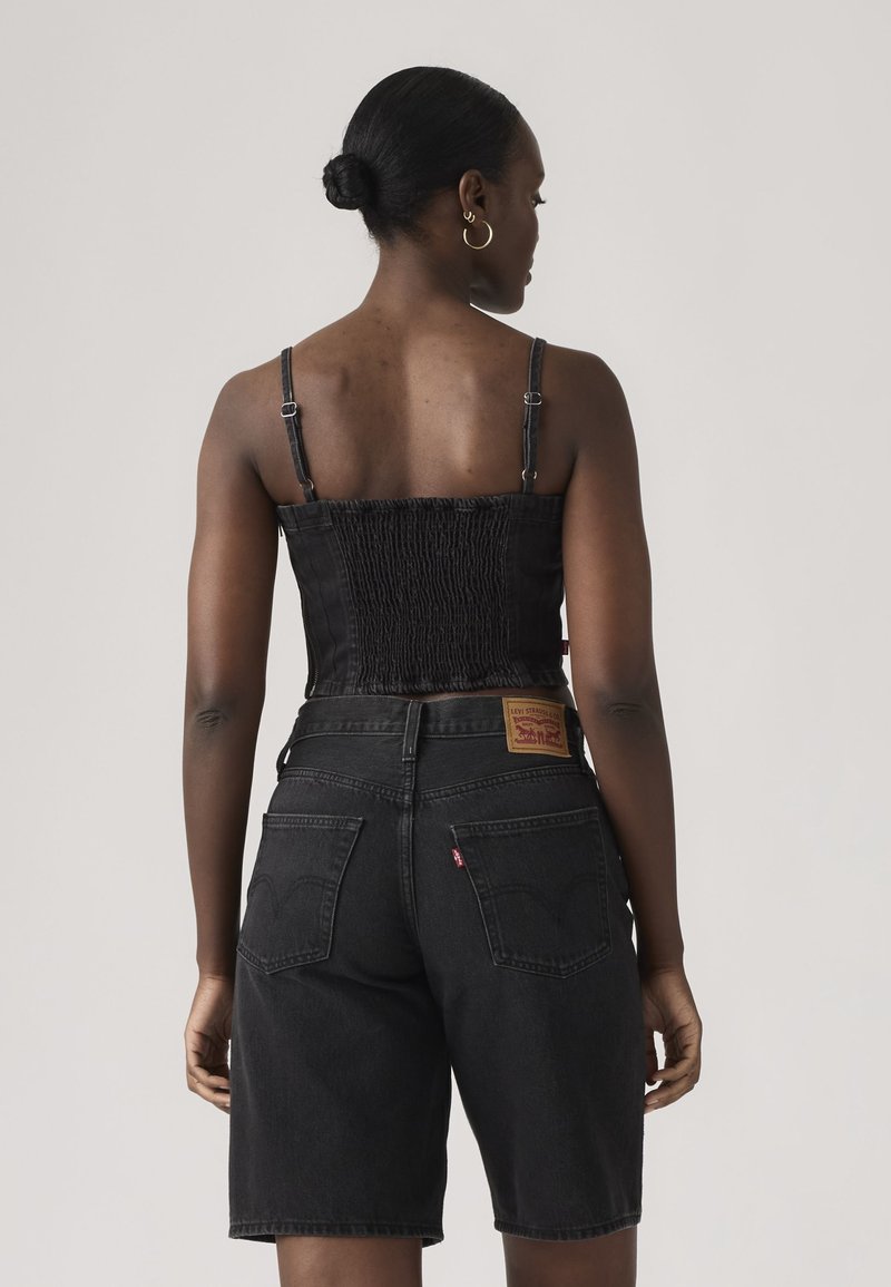 Femme aux cheveux attachés en chignon, portant un haut court smocké noir et un short en denim noir taille haute, vue de dos sur un fond neutre.