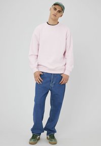 Sweatshirt en coton rose clair avec des poignets côtelés, associé à un jean en denim bleu ample et des baskets vertes. Design simple, coupe décontractée.