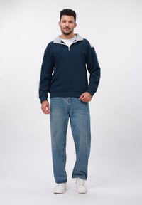 Uomo in piedi che indossa una felpa con zip navy blu e grigia, jeans azzurri chiari e sneakers bianche su uno sfondo bianco uniforme.