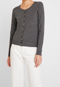 Kvinna iförd en åtsittande mörkgrå knappad kofta och högmidjade vita byxor, stående mot en enkel ljus bakgrund.