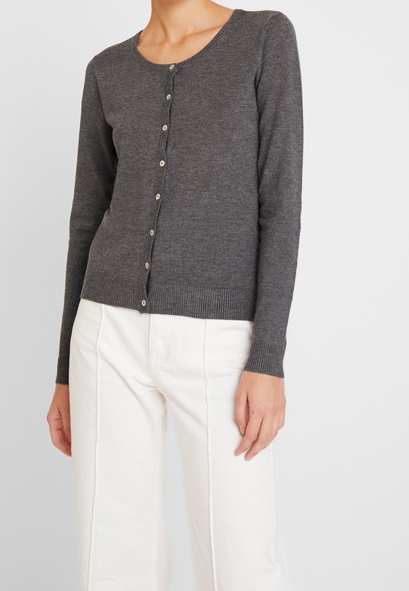 Kvinna iförd en åtsittande mörkgrå knappad kofta och högmidjade vita byxor, stående mot en enkel ljus bakgrund.