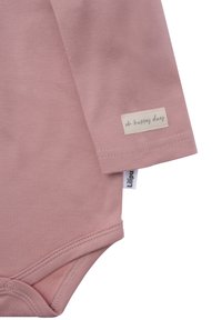 Hellrosa Baumwoll-Longsleeve-Bodysuit mit Druckknopfverschluss, versehen mit einem cremefarbenen Etikett, auf dem in schwarzer Schrift "oh happy day" steht. Geschmeidige Textur.
