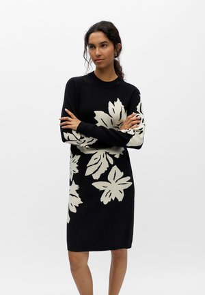 Femme debout, bras croisés, portant une robe noire longueur genou avec de grands motifs floraux blancs sur les manches et le corps.