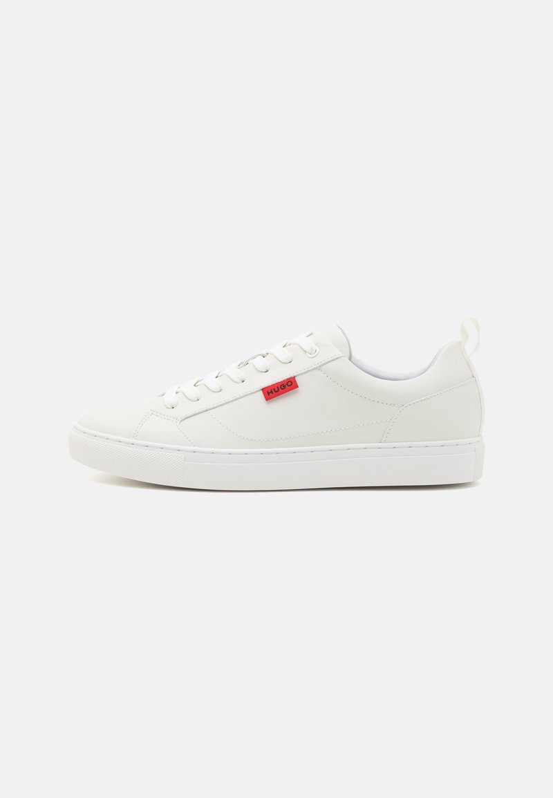 Baskets basses blanches avec des lacets blancs et une étiquette rouge indiquant "HUGO" sur le côté, présentées sur un fond clair uni.