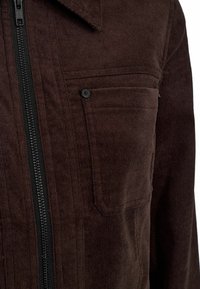 Rjav kordni jakna z črnim zadrgo spredaj, razširjenim ovratnikom in dvema žepoma na prsih s črnimi gumbi. Teksturovana tkanina in prilega kroj.