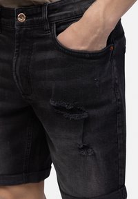 Person wearing black distressed denim shorts with rolled hems and hand inside front pocket.
