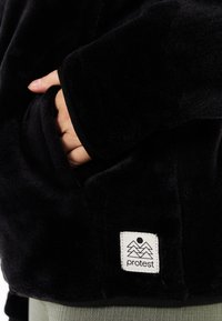 Schwarze Fleecejacke mit weicher Textur, auf der ein Hand in einer Tasche ist, sowie einem weißen Etikett unten rechts mit einem Berglogo.