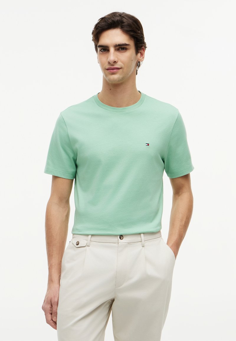 Jeune homme portant un t-shirt vert clair à manches courtes et un pantalon beige, debout avec une main dans la poche sur un fond blanc.