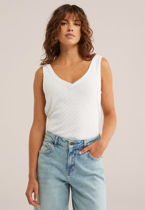 Donna con capelli castani ricci che indossa un top lavorato a maglia bianco senza maniche e jeans a vita alta color azzurro chiaro, mano nella tasca, sfondo neutro.