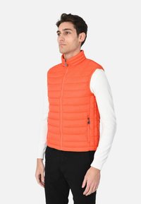 Gilet imbottito arancione con colletto alto, cuciture orizzontali e due tasche laterali con zip, indossato sopra una camicia bianca a maniche lunghe e pantaloni neri.