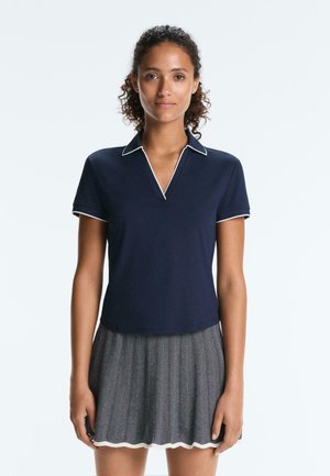 Jeune femme aux cheveux bouclés portant un polo bleu marine avec des bordures blanches et une jupe à motifs noirs et blancs, debout devant un fond uni.