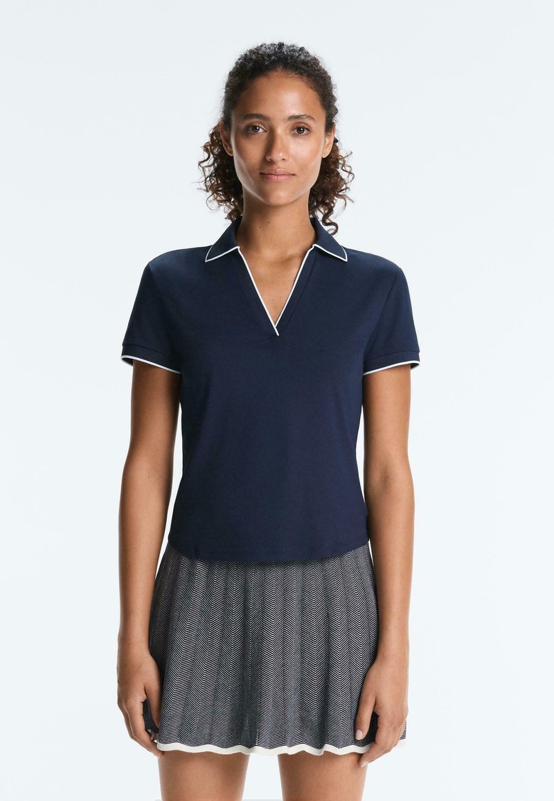 Mlada ženska s kodrastimi lasmi, oblečena v mornarsko modro polo majico z belim robom in črno-belo vzorčasto krilo, stoji pred enobarvnim ozadjem.