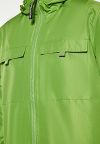 Giacca verde con zip, dotata di due tasche anteriori con patta e un'etichetta con la scritta "Urban Rain" vicino al cappuccio con cordino.
