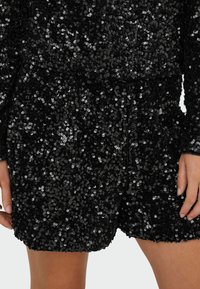 Sorte shorts i stof, prydet med sorte palietter og med en struktureret overflade. De har en snor i taljen og en afslappet pasform, der gør dem velegnede til forskellige anledninger.