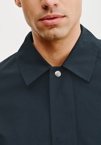 Ciemnoniebieska kurtka z klasycznym kołnierzem, srebrnym zatrzaskiem przy dekolcie i gładkim, teksturowanym wykończeniem tkaniny.