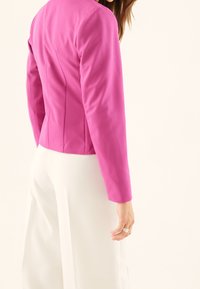 Dopasowany różowy żakiet z długimi rękawami i szwami z tyłu, zestawiony z białymi szerokimi spodniami, podkreślający gładką strukturę tkaniny.