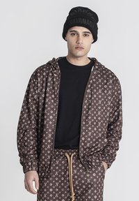 Brun zip-jacka med ett upprepande logomönster, elastiska ärmar och dragsko i midjan. Matcher med en svart tröja och en stickad mössa.
